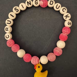 Beaded bracelet with pink cracked-agate and white beads, letter beads spelling CRUISING DUCKS, and a yellow rubber duck charm on a dark background.