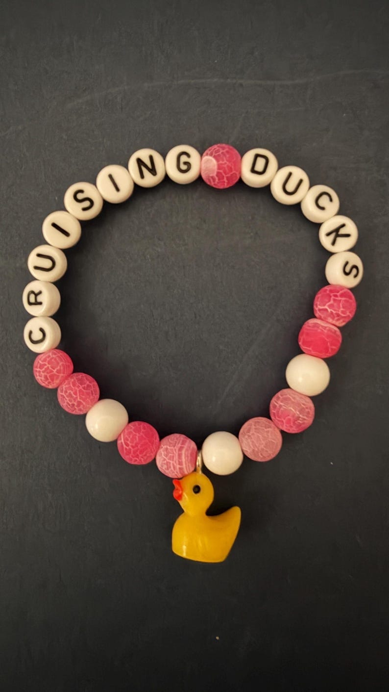 Beaded bracelet with pink cracked-agate and white beads, letter beads spelling CRUISING DUCKS, and a yellow rubber duck charm on a dark background.