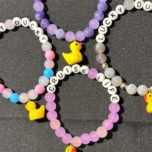 Four beaded bracelets with yellow duck charms and letter beads spelling CRUISE LIFE and LUCKY DUCK arranged on a dark surface.