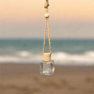 May include: A clear, square glass bottle with a wooden cap and a beige rope for hanging. The bottle is suspended against a blurred background of a beach and ocean, suggesting a car air freshener or decorative item.