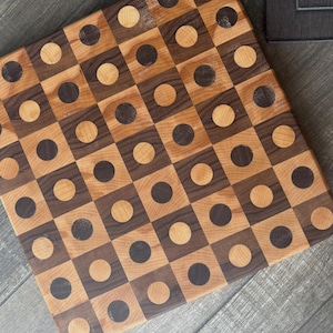 May include: A square wooden cutting board featuring a geometric pattern of alternating light and dark wood squares. Each square contains a contrasting circular inlay, creating a grid of light and dark circles across the entire surface.