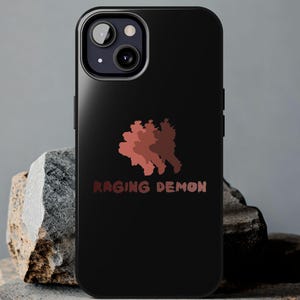 May include: Black phone case with a red and brown graphic of a stylized figure and the words "RAGING DEMON" in red. The phone case is displayed on a rough, gray stone surface.