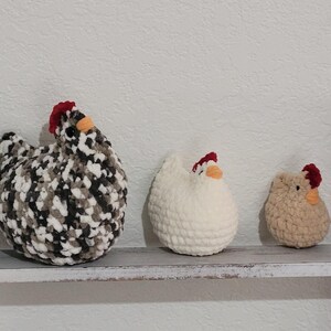 May include: Three handmade crochet chickens in varying sizes. The largest is speckled black, white, and gray. The middle chicken is white, and the smallest is tan. All have red combs and orange beaks, displayed on a rustic shelf.
