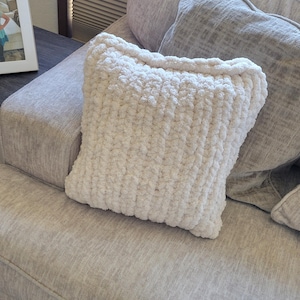 May include: A cream-colored, square pillow with a chunky knit texture rests on a light gray sofa. The pillow's soft, plush appearance is accentuated by the visible stitches. Other gray pillows are also on the sofa.