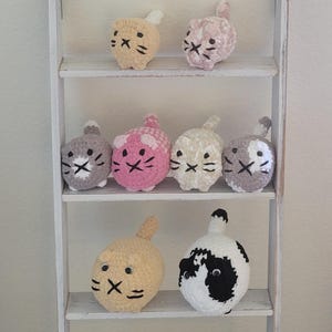 May include: A white wooden shelf unit displays nine crocheted cat-shaped toys. The cats are various colors, including yellow, pink, gray, and white, with black embroidered facial features. Each cat has a small tail.