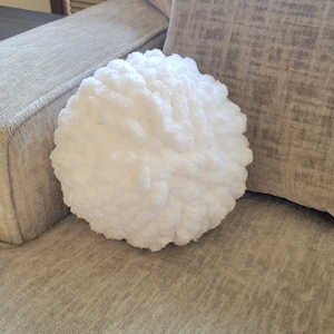 May include: A fluffy, white, round decorative pillow made of a soft, textured material. The pillow is placed on a gray upholstered sofa. The pillow's shape resembles a cloud or a ball of yarn.