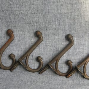 May include: Four antique bronze-colored metal coat hooks. Each hook has a curved design with a decorative top and a screw for mounting. The hooks are arranged in a row.