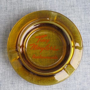 May include: Round, amber-colored glass ashtray with a circular design. The center features the text "Tony Roma's Restaurants" in red script. The ashtray has a smooth, glossy finish and a small indentation for resting cigarettes.