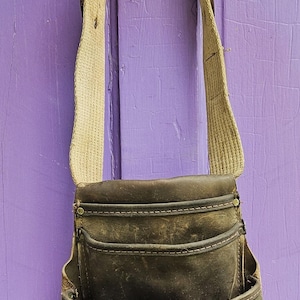 May include: A dark brown leather tool pouch featuring multiple pockets, a wide beige canvas shoulder strap with a metal buckle, and the text Weyerhaeuser printed in white on the front pocket. The pouch has visible stitching and metal rivets.