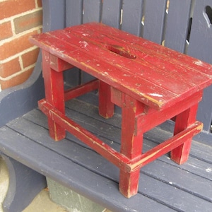 May include: A small, weathered red wooden step stool with a rectangular top and four legs. The stool has a hand-cut oval opening in the center of the top. The paint is chipped, giving it a rustic appearance.