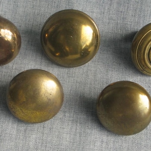 May include: A collection of five round brass door knobs with a metallic finish. Four of the knobs feature a smooth, plain surface, while one knob displays a distinct circular ring pattern around its center, all arranged in a group.