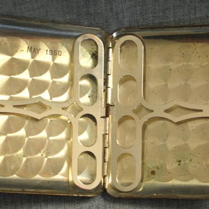 May include: Open, vintage gold-toned cigarette case. The interior features a decorative metal design and the inscription "MAY 1950". The case is rectangular with rounded corners and a hinged opening, showcasing a classic design.