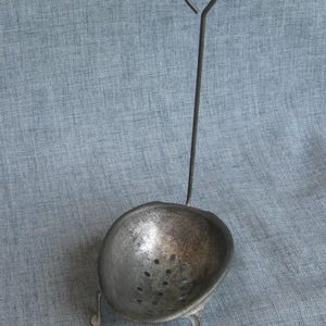 May include: A metallic tea strainer with a deep, oval-shaped bowl featuring small drainage holes. The strainer is supported by three short, sturdy legs and includes a long, slender vertical handle that terminates in a circular loop at the top.