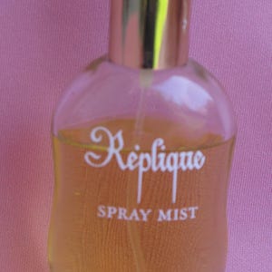 May include: A clear glass bottle of Réplique Spray Mist perfume with a gold-colored cap. The bottle has a rounded shape and contains a light amber liquid. The words "Réplique", "Spray Mist", and "Raphael" are printed on the bottle in white.