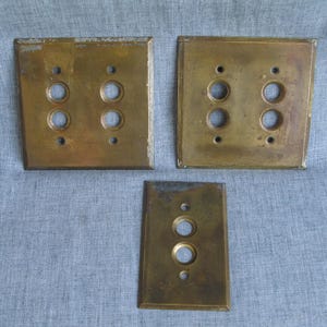 May include: Three rectangular brass switch plates with a weathered, tarnished finish. Two plates are square with four circular openings for switches and four screw holes. One plate is narrow with two circular openings and two screw holes.