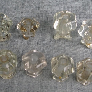 May include: A set of eight clear, faceted glass knobs arranged in two rows. Each knob features a hexagonal shape with a central circular opening and a smooth, reflective surface. The knobs are transparent and display a geometric, multi-sided design.