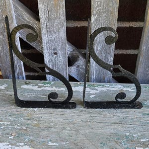 May include: Two black metal shelf brackets featuring an S-shaped scroll design with a hammered texture. Each bracket has a flat mounting plate at the top and a flat base, with decorative curled ends at the top and bottom of the curved support arm.