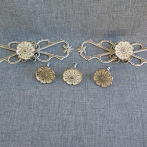 May include: Two elongated, off-white and metallic gold decorative drawer pulls with intricate scrollwork and a central floral rosette, accompanied by three matching circular floral knobs, each featuring a central screw for mounting purposes.