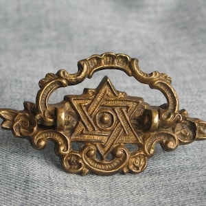 May include: A brass drawer pull featuring a central six-pointed star design with a circular center point. The hardware has an ornate, decorative border with scrolling leaf-like details and a hinged, curved handle attached to the top section.