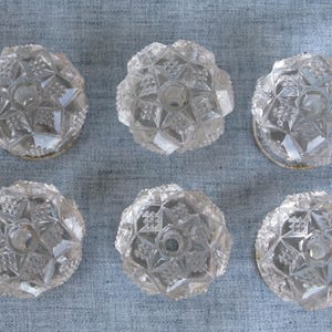 May include: A set of six clear glass knobs, each featuring a circular shape with a faceted, star-like geometric pattern and a central hole for mounting. The glass is transparent with sharp, angular edges that catch the light across the surface.