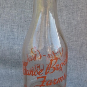 May include: Clear glass milk bottle with orange lettering. The bottle has a narrow neck and a rounded body. The text on the bottle reads "Canoe Brook Farms, Summit, N.J."
