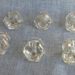 May include: Six clear, faceted glass cabinet knobs with a hexagonal shape. The knobs have a vintage aesthetic and are arranged on a gray fabric surface. The knobs are likely for furniture restoration or decorative purposes.