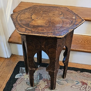 May include: A hexagonal wooden side table with a dark brown finish, featuring intricate carved patterns on the top surface and legs. The tabletop displays a central circular motif with detailed engravings, while the legs have arched cutouts.