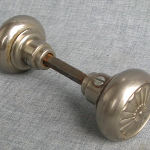 May include: A pair of silver-toned metal door knobs connected by a square-shaped metal spindle. Each knob features a smooth, rounded surface with a decorative sunburst or fan-like pattern embossed on the face and a small screw on the side.