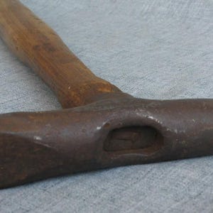 May include: A metal hammer featuring a dark, weathered iron head with a wide, flat striking surface on one side and a pointed, chisel-like end on the other. The tool includes a long, cylindrical wooden handle with a natural brown grain finish.