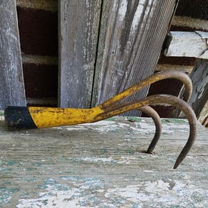 May include: A hand cultivator with three curved metal tines and a yellow handle featuring a black end cap. The metal shows signs of oxidation and wear, with patches of rust visible along the tines and the yellow painted handle surface.