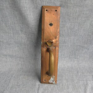 May include: A vintage copper door handle with a brass handle. The rectangular plate has a tarnished finish with signs of wear and tear. The handle is curved and has a classic design, with a keyhole and mounting holes.