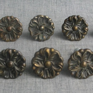 May include: A set of ten round metal cabinet knobs featuring a dark bronze finish and a raised floral relief pattern. Each knob has a central circular protrusion and a series of curved, petal-like segments radiating outward from the center.