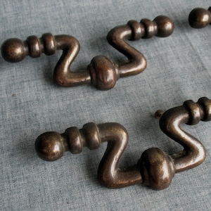 May include: Close-up of several antique bronze-colored drawer pulls. Each pull has a distinctive S-shaped design with a ball-shaped end and a central, rounded feature. The pulls are arranged on a textured, blue-gray surface.