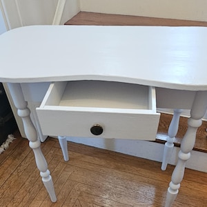May include: A white painted wooden vanity table featuring a smooth, curved tabletop and a single central drawer. The drawer is equipped with a small, round, dark-colored knob. The table is supported by four slender, turned legs with a decorative spindle design.