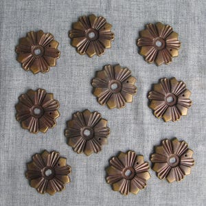 May include: Nine antique brass-toned flower-shaped decorative hardware pieces. Each piece has a central hole and radiating petal-like designs. The hardware is arranged on a gray fabric surface.