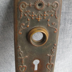 May include: Antique bronze door plate with ornate floral and scrollwork detailing. Features a keyhole and a circular opening for the doorknob. The plate has a rectangular shape with rounded edges and a weathered appearance.