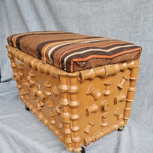 May include: A brown storage bench with a bamboo-style design and a striped cushion. The bench has a rectangular shape, is mounted on small wheels, and features decorative spool-shaped accents. The cushion has brown, orange, and white stripes.