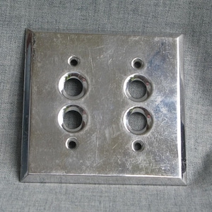 May include: A square, brushed-metal light switch plate with four circular openings for switches and two smaller holes for screws. The plate has a slightly beveled edge and a reflective surface.