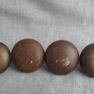 May include: Four weathered, spherical metal balls in varying shades of brown and bronze are arranged in a row. The balls have a rustic, aged appearance, suggesting they may be antique or vintage items.