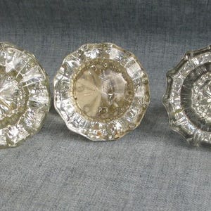 May include: Five round glass door knobs arranged in a horizontal row. Each knob features a faceted, starburst-like pattern radiating from the center toward the scalloped edges. The glass varies in clarity, ranging from clear to light amber tones.