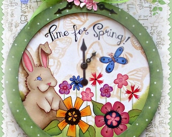 Op de afbeelding: Een decoratieve klok met een groen frame en een lentethema. De wijzerplaat toont een konijn, bloemen en een vlinder. De tekst "Time for Spring!" staat op de wijzerplaat. Ontworpen en geschilderd door Sharon Bond (c) 2015.