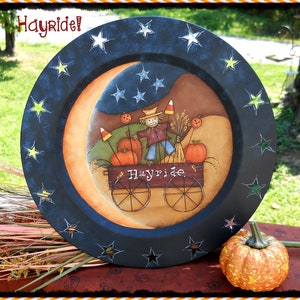 Puede incluir: Un plato pintado de negro con un diseño de luna creciente y estrellas. El plato presenta una escena pintada de un espantapájaros montando un paseo en un carro de heno con calabazas y maíz. El texto "Hayride!" está escrito en el plato.