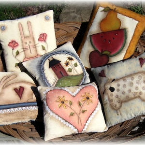 May include: A collection of six handmade decorative cushions with various designs, including a bunny, a house, a heart, a sheep, and fruit. The cushions are made of fabric and are decorated with paint and stitching. The cushions are arranged in a wicker basket.