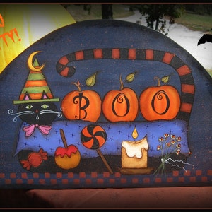 May include: A black cat wearing a witch's hat sits on a blue blanket with three orange pumpkins that spell out "BOO" and a lit candle. There is a candy cane, an apple, and a candy on the blanket.