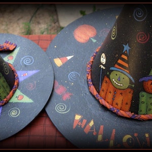May include: Two black witch hats with orange and green painted faces and a star and swirl design. The larger hat has the word "Halloween" written on it.