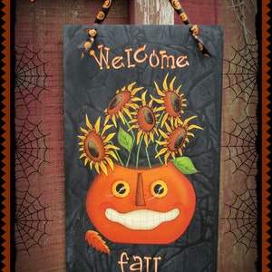 May include: A black painted wooden sign with the text "Welcome fall!" and a jack-o'-lantern with sunflowers coming out of the top. The sign is hanging on a wooden door with a spiderweb in the background.