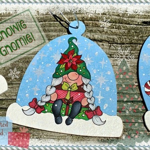 May include: Three wooden Christmas baubles shaped like snow globes, each featuring a different gnome design. The first gnome is holding a small Christmas tree, the second gnome is sitting with a poinsettia flower, and the third gnome is holding a candy cane with a cardinal bird perched on its hat. The baubles are painted in bright colours and have a whimsical, festive feel. The text "Snowie Gnome!" is visible at the top of the image.