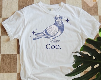 Puede incluir: Camiseta blanca con una ilustración azul de una paloma con gafas de sol y la palabra "Coo." debajo. El diseño incluye estrellas. La camiseta está hecha de un material suave y cómodo.