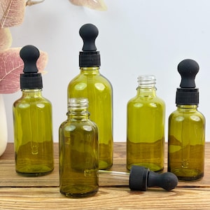 May include: Several olive-green glass dropper bottles of varying sizes, each with a black rubber bulb and a black screw-on cap. One bottle has the dropper detached. The bottles are arranged on a wooden surface.