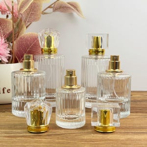 Puede incluir: Una colección de frascos de perfume de vidrio transparente con pulverizadores dorados y tapones decorativos. Los frascos tienen un diseño acanalado y están dispuestos sobre una superficie de madera. Algunos frascos tienen un tapón cuadrado o facetado.